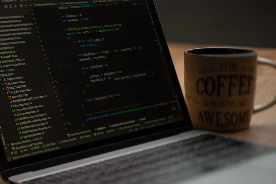 A close-up of a laptop displaying lines of computer code, with a yellow ceramic mug sitting beside it on a wooden surface. The mug has a humorous message about coffee, reading 'This coffee is making m