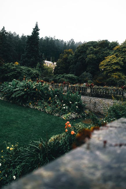 A scenic outdoor garden landscape featuring a lush green lawn bordered by flower beds filled with colorful blooms and foliage. The garden is set against a background of dense, mature trees and a hills