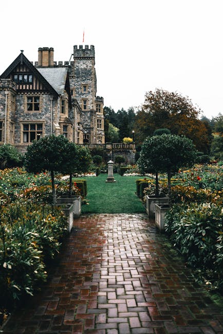 A scenic outdoor garden landscape featuring a lush green lawn bordered by flower beds filled with colorful blooms and foliage. The garden is set against a background of dense, mature trees and a hills
